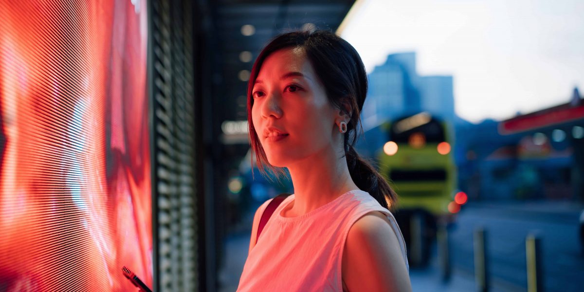 Confident young Asian businesswoman standing against illuminated LED digital display in the city, lit by red neon coloured lights, using smartphone in downtown city street in the evening. Lifestyle and technology
